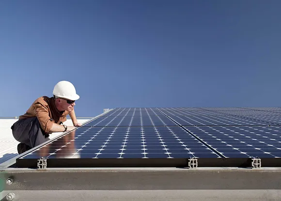 A person in a white hard hat and brown jacket inspecting solar panels on a rooftop, with a clear blue sky in the background.