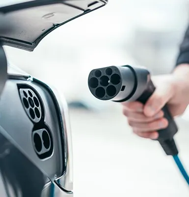 A close-up of a white electric vehicle being charged, with a charging cable connected to the car's charging port, greenery and a building in the background.