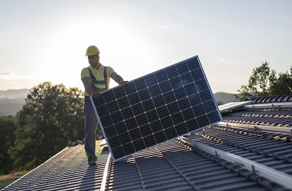 Single worker carrying a solar panel on a rooftop during sunset, representing commercial solar financing solutions in Denver, CO.