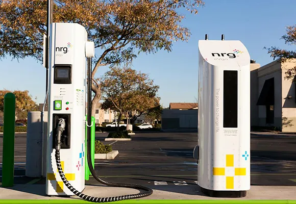 Public electric vehicle charging stations with white and green signage situated in a parking lot with trees in the background.