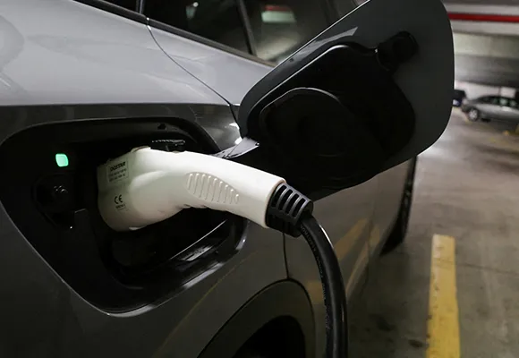 A white electric vehicle being charged at a charging station, with a close-up of the charging cable connected to the car's charging port.