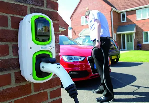 A red electric vehicle parked near a charging station mounted on a brick wall, with greenery in the background.