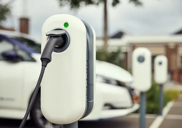 A white electric vehicle parked in a driveway, with a charging station mounted on a wooden fence nearby.