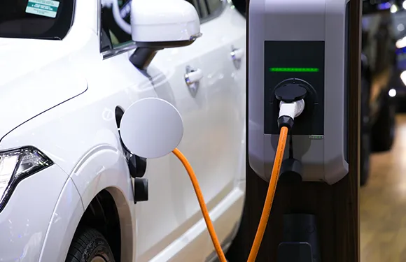 A white electric vehicle parked in a driveway, with a charging station mounted on a wooden fence nearby.