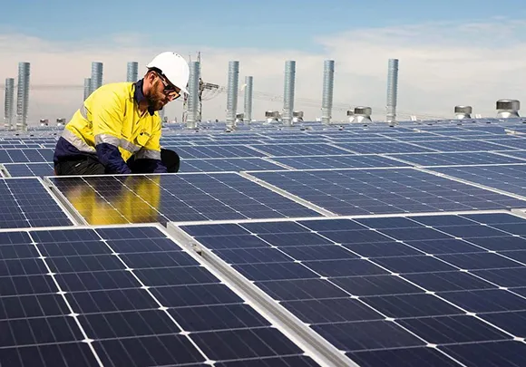 Worker wearing a yellow safety jacket inspecting or cleaning large solar panels on a rooftop.