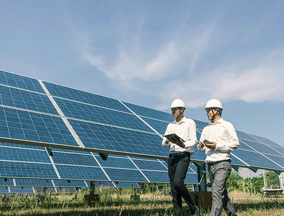 Two people in white hard hats standing in front of a solar panel array under a clear blue sky, with one person pointing at the panels.